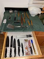 Full view of various kitchen utensils including ladle, masher, grilling tools with wooden handles, colander and spatula arranged on a green cloth.