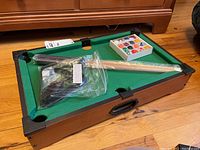 Miniature pool table with green felt surface and brown frame with corner pockets. Two wrapped wooden pool sticks, black triangle rack and box of pool balls on table.