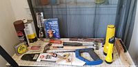 Photo showing various hand tools including a saw with blue handle, yellow flashlight, tape measure, hammer, pry bar, and spray can.