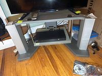Overall front view of gray TV stand with storage shelving, flat top surface holding a TV, and glass middle shelf supporting a media device.