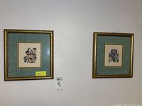 Two framed floral prints hanging on a wall, each with greenish-blue mats and gold decorative frames, side by side.