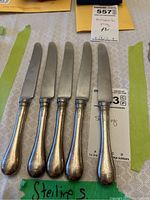Five sterling silver cutting knives displayed parallel with rounded, slightly worn handles and shiny blades on a neutral patterned cloth.