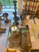 Seven-piece brass lot on white table including mailbox, candleholders, coal pot, and serving dish near window with curtain