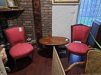 Pair of red upholstered wood frame chairs positioned around a round wooden table on dark hardwood floor with brick and stucco walls in background.