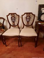 Pair of ornate wood frame chairs with upholstered light beige seats, placed side by side on wooden floor, showing decorative carved backs with gold accents.