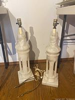 Pair of white carved stone base table lamps standing on a wood floor against a white wall. Both lamps have no shades and show electrical cords and sockets at top wrapped partially in bubble wrap.