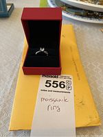 Moissanite ring in a red jewelry box atop a yellow envelope with the lot number and handwritten note indicating 'moissanite ring'.