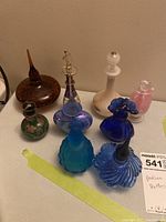 Nine assorted vintage glass perfume bottles displayed on a flat white surface, showing various shapes, colors and intricate stopper designs.