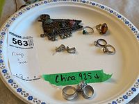 Full lot on a white plate with blue flower rim, showing brooch, rings, and earrings, highlighting two rings marked China 925 on green tape