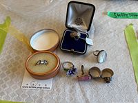Seven sterling silver rings displayed on a tablecloth, including rings with gemstone settings and intricate silver designs; some rings are in cases.