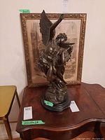 Front view of full statue on wood table with framed artwork behind, green tape marking height.