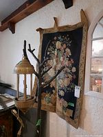Full view of floral tapestry hanging on wall with base of wooden coat tree and brass hanging lantern nearby for size context.