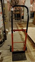 Front view of hand cart upright with orange lower frame and black handlebar, flat loading plate visible.