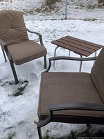 Two metal lawn chairs with beige cushions set outdoors in a snowy lawn environment alongside a small side table with brown wooden slats and metal legs.