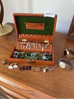 Full view of brown jewelry case with green velvet lining showing arranged tie tacks, cuff links, and two watches positioned nearby