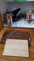 Cabinet shelf with two black speckled Heritage rock pots and a tall stainless steel stock pot with lids, wooden cutting board on counter in front