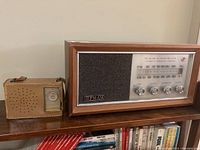 Photo showing the Emerson transistor radio in its tan leather case next to the Philips FM AM radio in a wooden cabinet, both displayed on a wooden shelf.