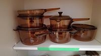 Overview of six amber tinted glass pots with lids on white shelving, showing various sizes and handle styles.