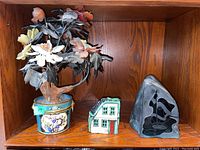 Three items displayed on wooden shelf including faux floral arrangement in pot, painted house figurine, Inuit carving