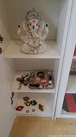 Full shelf view showing the white Ganesha statue on top shelf, assorted wristwatches and watch parts on the middle shelf, and bracelets with watch parts on the bottom shelf.