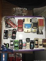 Top view of 18 assorted toy vehicles including cars, trucks, and a helicopter, displayed on a white tray with purple edges.