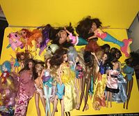 Top view showing diverse assortment of dolls laid out against a yellow background, including Barbie dolls, a Dora doll, miniature dolls, and several My Little Pony figures.