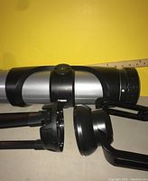 Side view of silver and black kids telescope tube alongside folded black tripod with yellow background and ruler for scale.