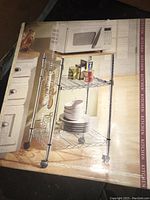 Box with image of assembled microwave cart showing chrome metal frame with two wire shelves, sliding shelf and metal hooks.