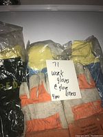 Four gloves total: one pair of used heavy-duty work gloves with orange cuffs and leather palms alongside three pairs of new Firecraft Gore-Tex glove liners in plastic packaging, two blue and one black pair visible.