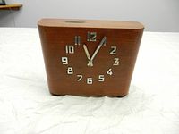 Front view of wooden clock with metal numbers and hands showing the time 12:03.