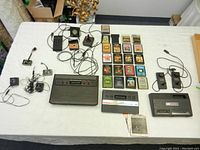 Complete overview of the three consoles: Atari 2600 (black with woodgrain), Atari 2600 Junior (black with silver stripes), and Coleco Gemini (black). Includes 17 game cartridges laid out, several controllers including joystick and numeric keypad, and cables.