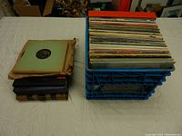 Wide shot of blue plastic crate packed with over 50 vinyl records and stack of 78 RPM records with three books on table.