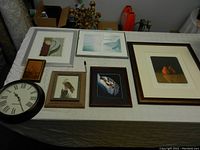 Several framed prints, a watercolour, feather art, a signed print, and a round wall clock arranged on a table.