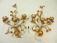 Photo showing front view of pair of metal leafy gold-tone wall sconces with floral details, placed on white surface alongside a marker for scale.