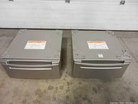 Front and top view of two metal pedestal risers with storage drawers closed, placed side by side on a concrete floor.