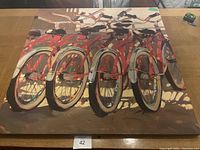 Front view of a large painting showing multiple red bicycles arranged closely together, focusing on the wheels and handlebars, laid flat on a wooden table.