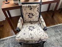 Front view of one bergere chair showing floral fabric with black flowers on beige background, black painted wood frame with carved detail.