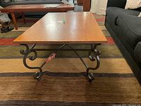 Front view of rectangular coffee table with maple wood top and decorative black metal scroll base, on a multicolored rug in front of a dark couch.