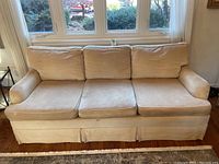Front view of beige fabric couch showing three seat cushions and three back cushions, set against large windows.