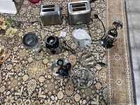 Full view of all kitchen appliances and cookware arranged on patterned carpet. Includes two toasters, blender parts, manual ice crusher, egg poacher, kettle, saucepan with lid.