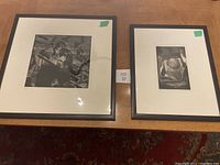 Two framed lithographs on a wooden table, each with white matting and black frames, showing black and white abstract and figurative designs.