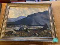 Large framed oil painting of mountainous landscape with river and grassy foreground under cloudy sky.