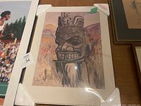 Poster featuring a carved totem pole with mountains and trees in the background, protected in plastic wrap with corner protectors.