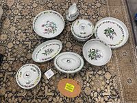 Porcelain serving dishes arranged on carpet, showing floral pattern on white base, various sizes and shapes with lids and some uncovered.