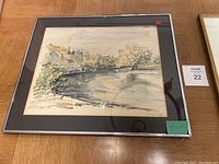 Framed watercolour river landscape painting with black frame and glass, signed, placed on wooden surface.