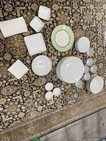 Full view of white china dinnerware including plates, small dishes, cups, saucers, and serving pieces arranged on patterned rug.