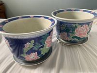 Two large ceramic planter pots with matching saucers, placed on fabric, showing floral designs and no damage.