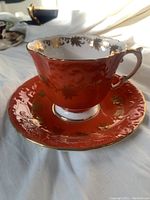 Orange Aynsley tea cup and saucer set with gold floral patterns and trim, showing the front and side of the tea cup and matching saucer.