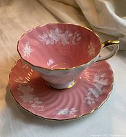 Photo of the pink Aynsley tea cup with white floral pattern and gold trim on the handle and rim, placed on matching saucer with similar floral and swirl designs.