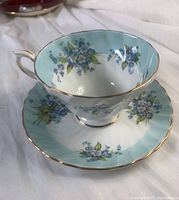 Front view of tea cup placed on saucer showing Forget Me Not blue flowers, swirl light blue background band and gold trim edges
